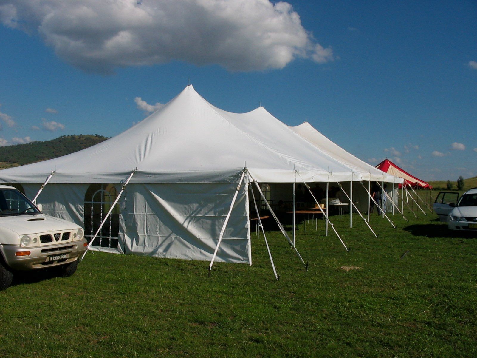 A large, white event tent set up on a grassy field under a sunny blue sky, with vehicles parked nearby.