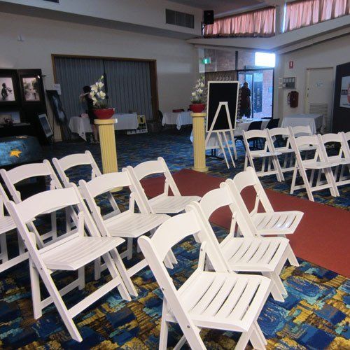 Rows of white folding chairs set up in an indoor hall, with a red carpet aisle, floral pedestals, and tables in the back.