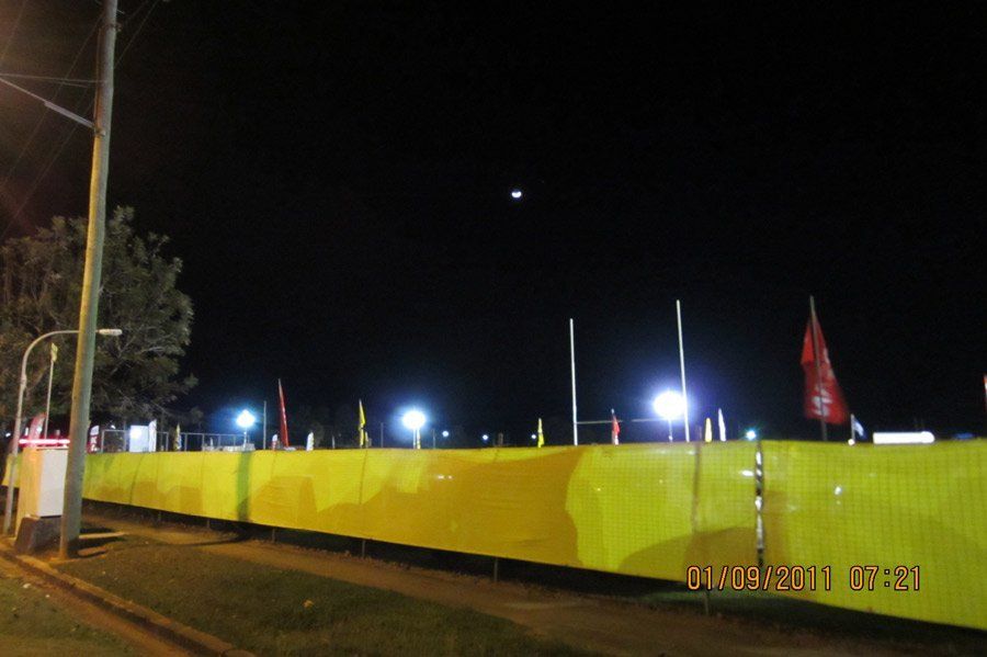 A long yellow construction fence lines a street at night, illuminated by bright artificial lights under a dark sky.