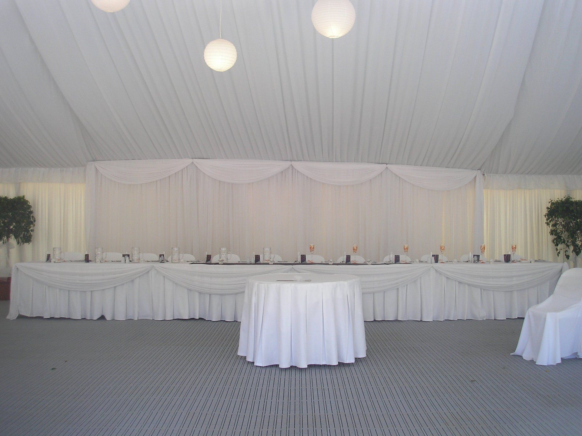 A long table with white floor-length tablecloths, draped swag, and a small round table in a white-draped event tent.