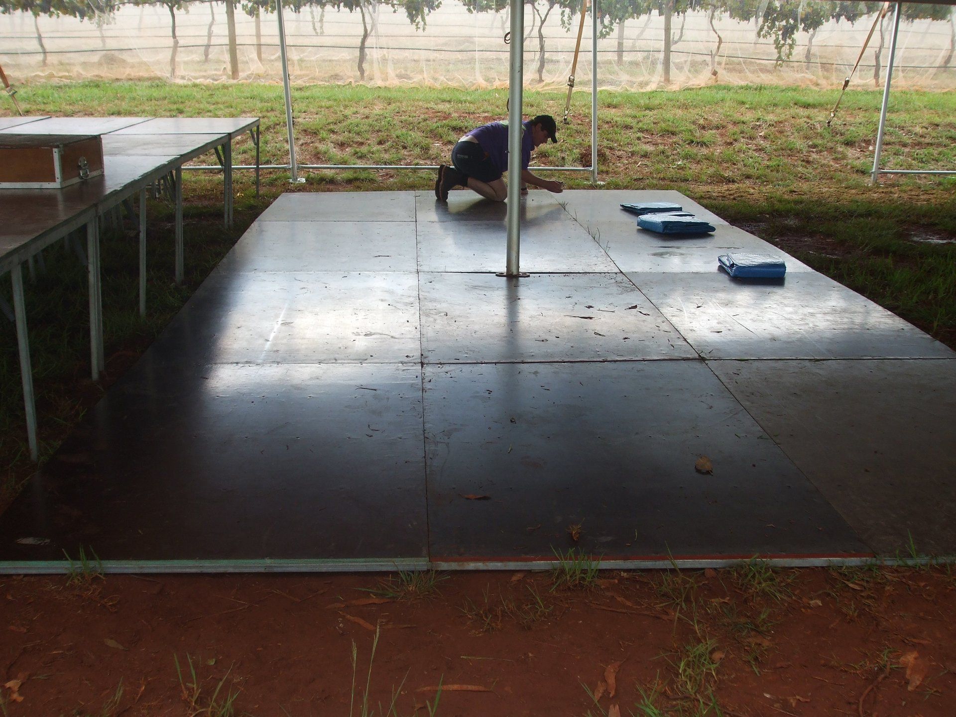 A person assembles modular wooden floor panels under a large outdoor tent frame on a grassy field.