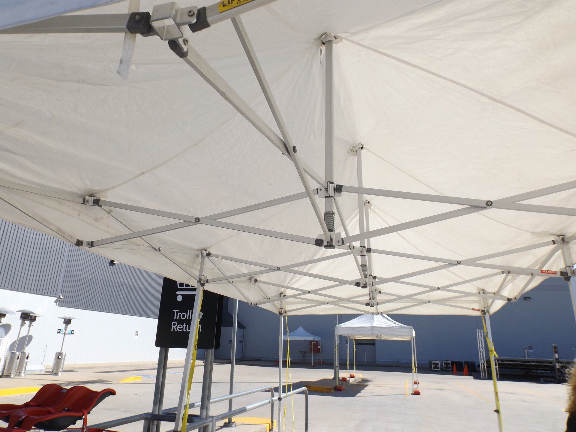 View from beneath a white pop-up canopy tent showing its metallic structural frame against an outdoor lot.