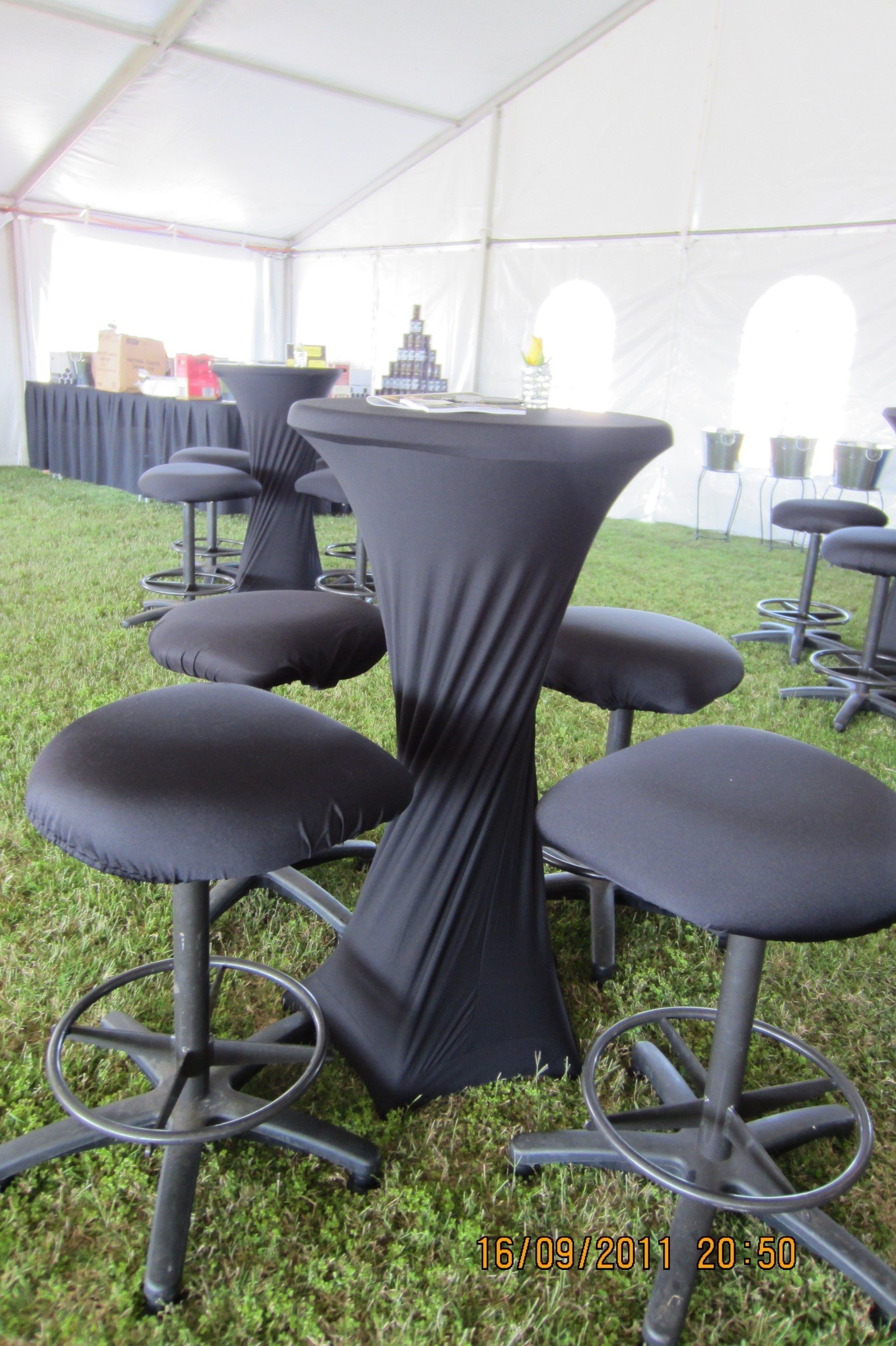 High-top tables with black fitted covers and matching bar stools set up on grass under a large white event tent.