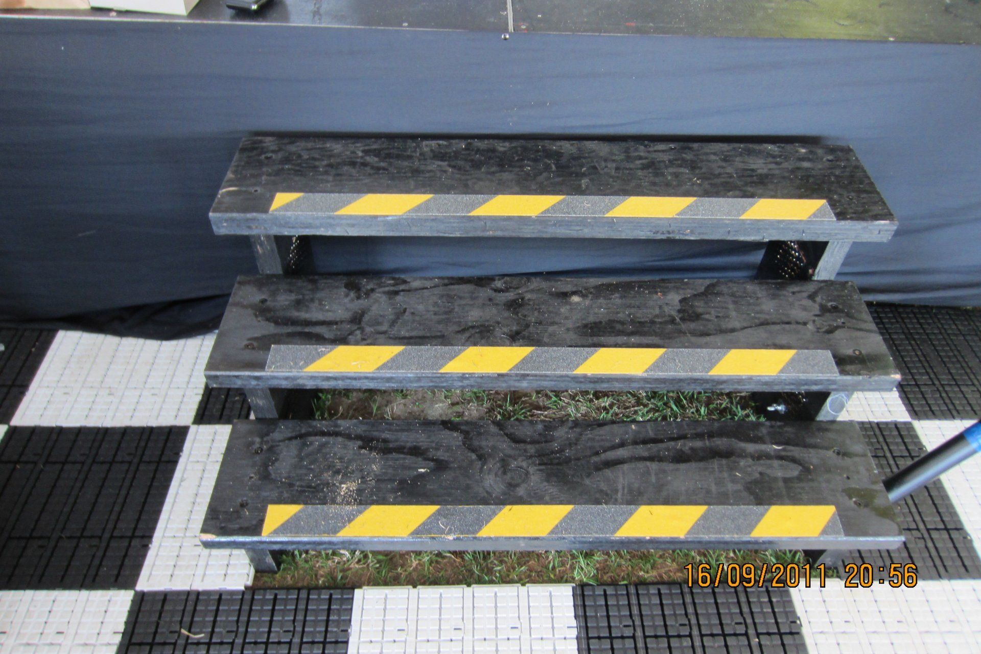 A three-step portable black staircase with yellow caution stripes on the edge of each step, set on a checkered floor.