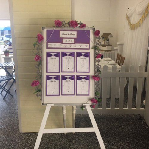 A white easel displaying a purple and white wedding seating chart adorned with pink roses, set in a light-colored room.