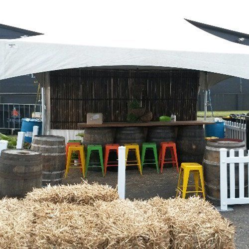 A rustic outdoor bar under a white tent, featuring wooden barrel stools, colorful metal chairs, and straw bale seating.