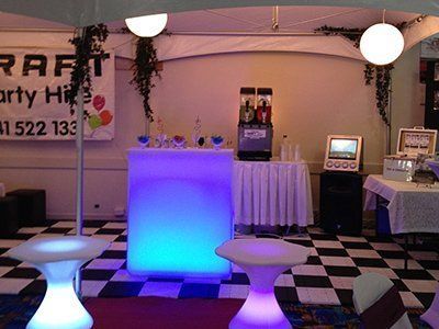 An event setup with an illuminated blue bar, two glowing cocktail tables, a slushie machine, and a checkered floor.