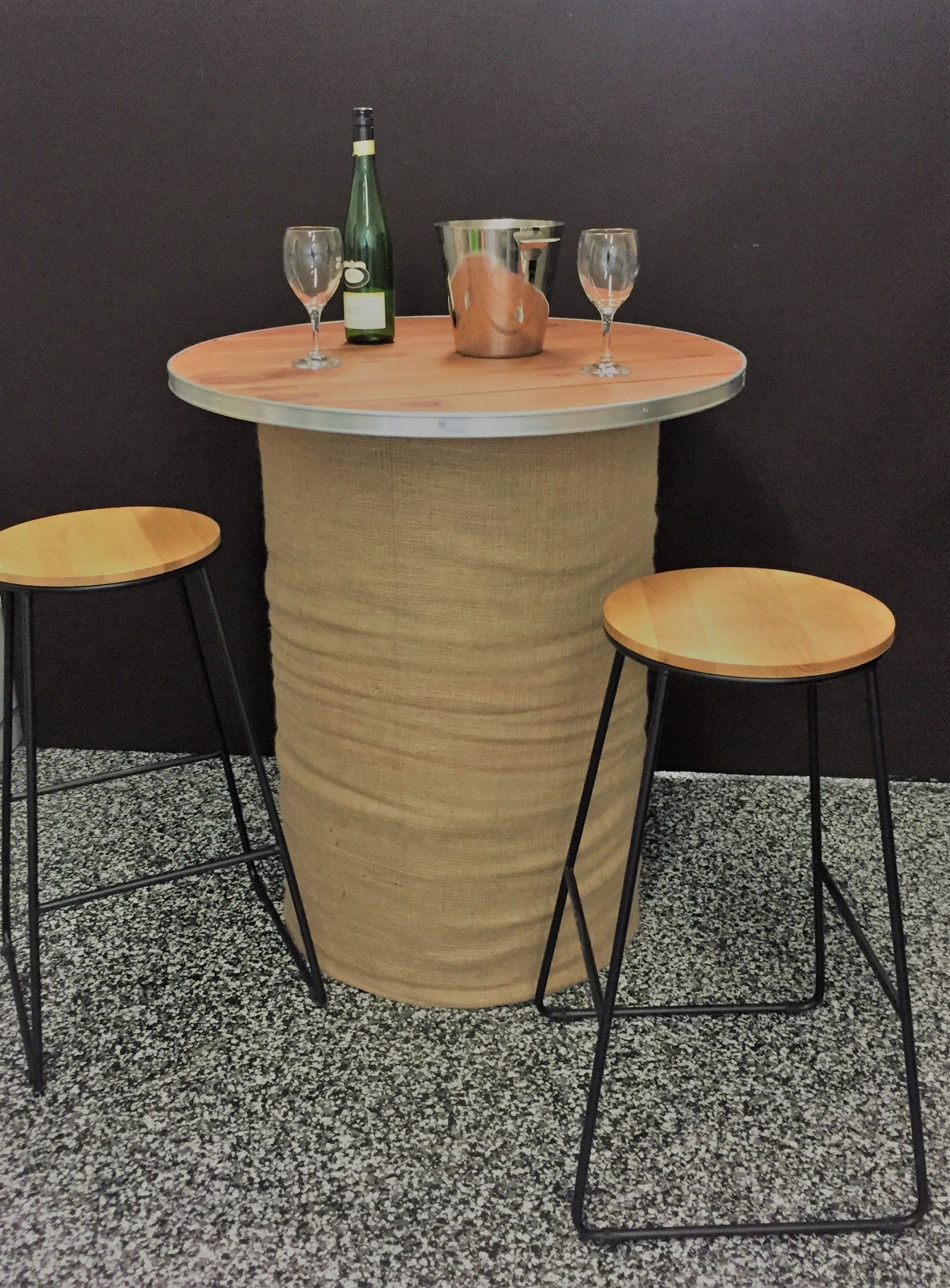 A burlap-covered barrel table topped with wine glasses, a bottle, and a metal bucket, flanked by two stools.