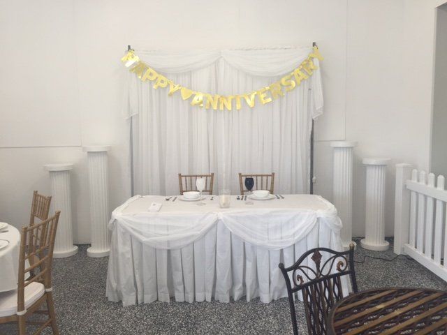 A decorated sweetheart table for an anniversary with white linens, chairs, and a gold 