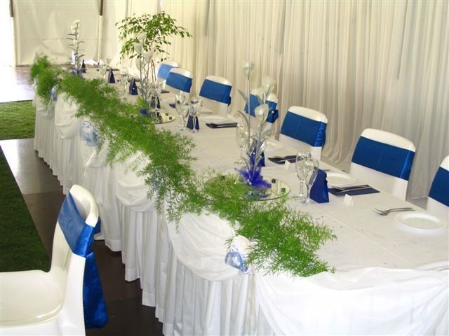 A long, banquet-style table set for an event with white tablecloths, chairs with blue sashes, and a green vine runner.