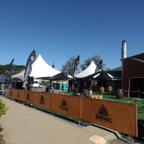 Bundaberg-branded wooden barriers stand before white event tents and a patio area on a clear, sunny day.