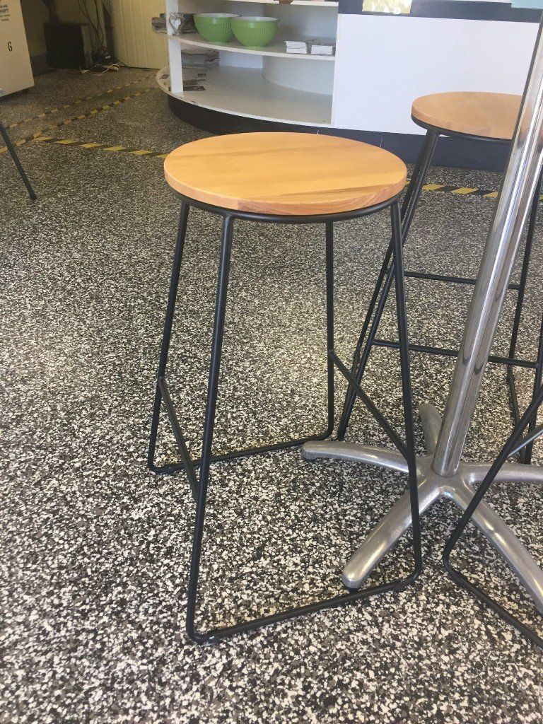 A metal-frame bar stool with a light wood seat, standing on a speckled floor next to a table leg.