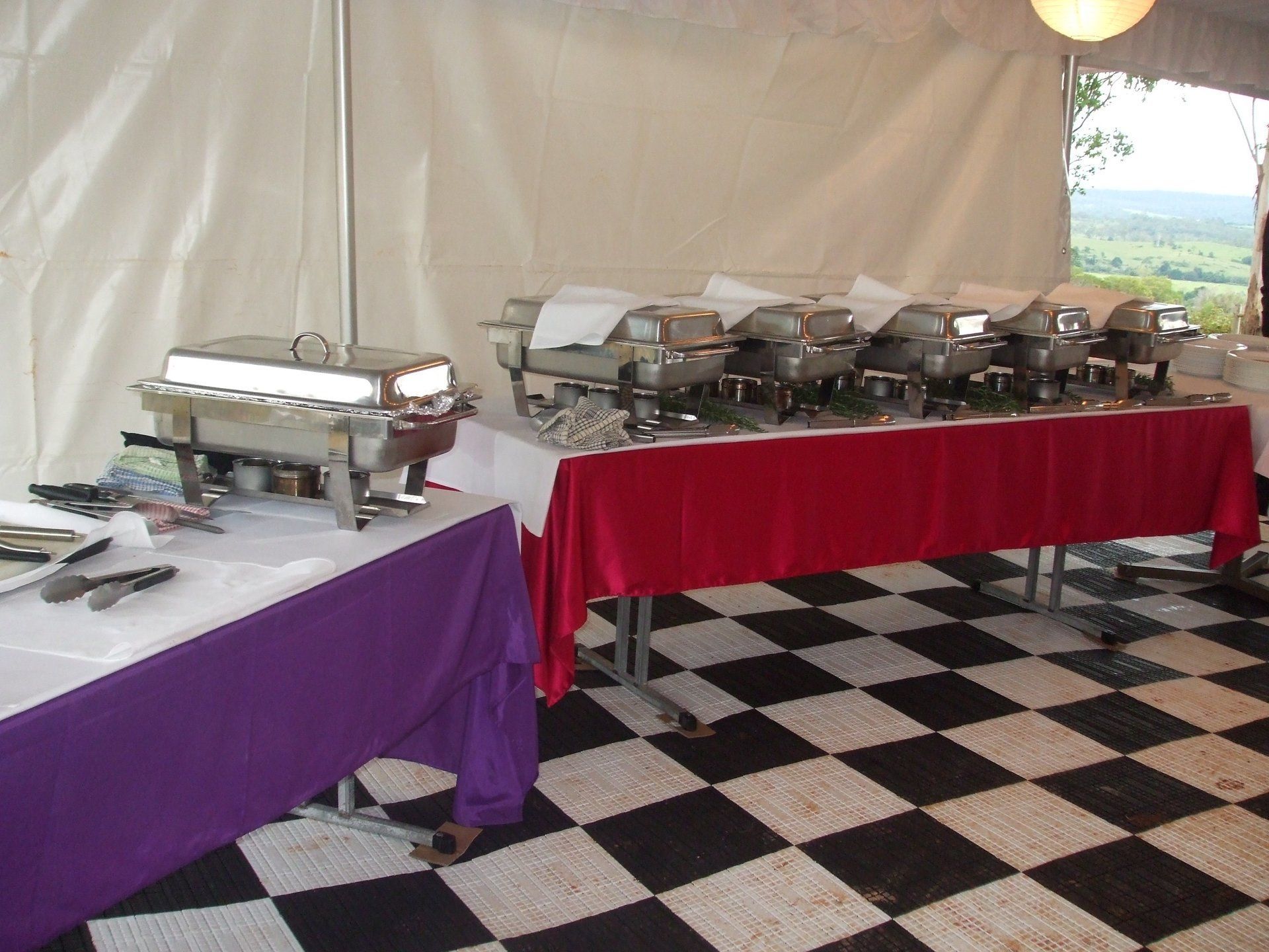 A buffet setup with stainless steel chafing dishes on tables with purple and red cloths on a checkered floor inside a tent.