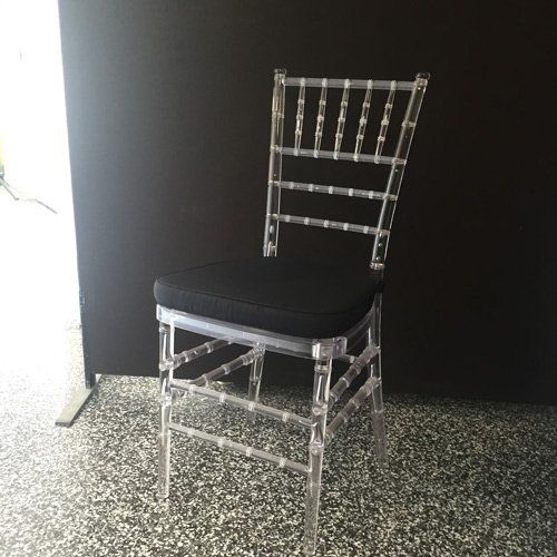 A clear, acrylic Chiavari chair with a black cushion, standing on a speckled floor against a dark background.