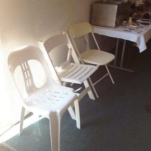 Three white plastic chairs lined up in a room next to a table with a white tablecloth.