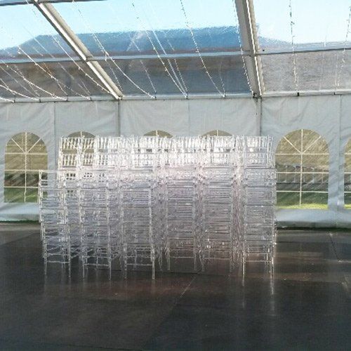 A large stack of clear, ghost-style chairs sits in the center of a white event tent with arched windows.