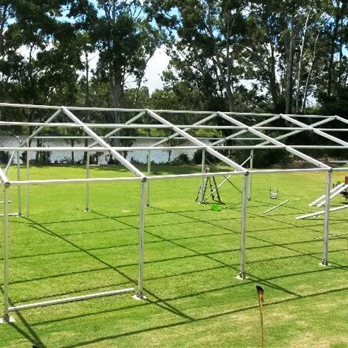 The metal frame of a large rectangular marquee tent stands on a lush green lawn near trees and a body of water.