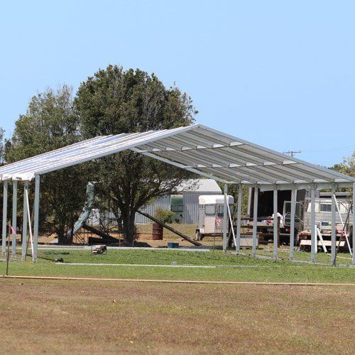 A metal frame structure for a carport or building stands in a grassy field on a sunny day with trees in the background.