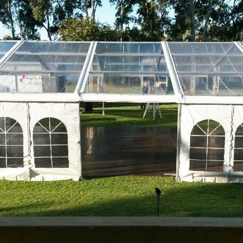 A large white frame marquee tent with arched windows and a transparent roof stands on a grassy lawn.