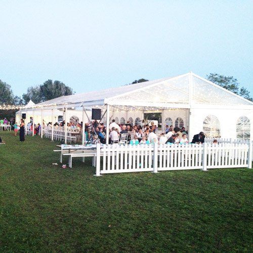 A white tented event on a grassy field, enclosed by a white picket fence, with people seated at tables inside.
