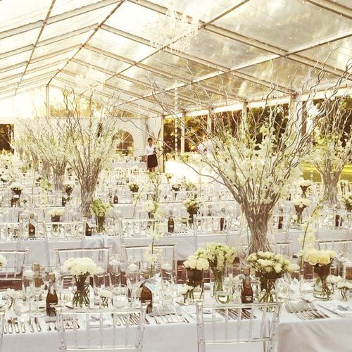 Elegant tables in a transparent marquee tent, decorated with tall white floral centerpieces and clear chairs.