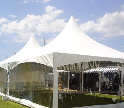 Two white high-peak event tents with clear plastic walls stand on a grassy lawn under a blue, cloudy sky.