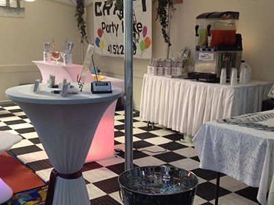 A party setup with a black-and-white checkered floor, white cocktail tables, and a refreshment station with slushies.