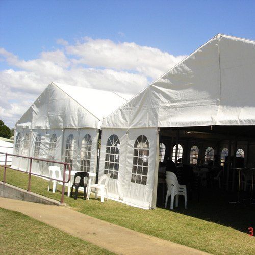 Two large white event tents with arched windows stand on a green lawn beside a paved walkway under a blue sky.