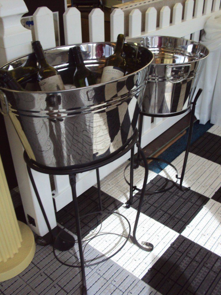 Two large, silver metal ice buckets on black metal stands holding wine bottles, situated on a black and white tiled floor.