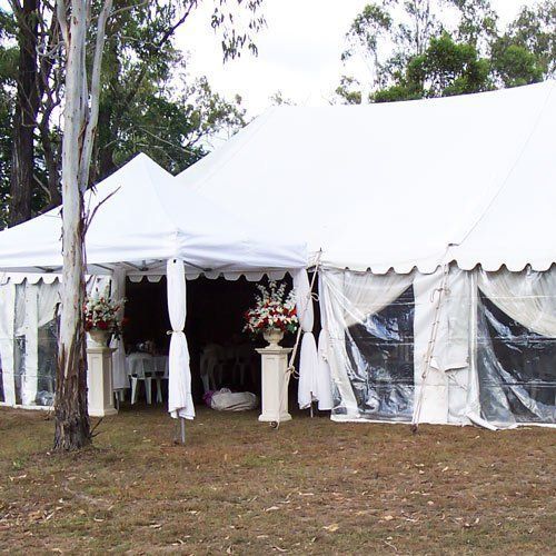 A large white event tent with clear plastic sides, adorned with floral arrangements, set in a wooded outdoor area.