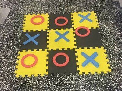 A tic-tac-toe game made of yellow and black foam tiles with red 'O' and blue 'X' pieces on a speckled surface.