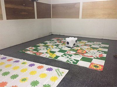 A floor-level shot of a Snakes and Ladders mat with a large soft die and a Twister mat on a grey floor in a room.