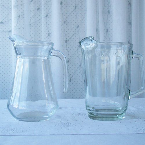 Two clear glass pitchers sit side-by-side on a white surface against a white curtained background.