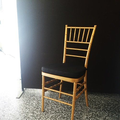 A bamboo-style wooden Chiavari chair with a black cushion sits on a speckled floor against a solid black backdrop.