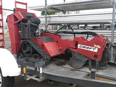 A red Craft stump grinder on tracks sits on a flatbed trailer outdoors next to metal scaffolding.