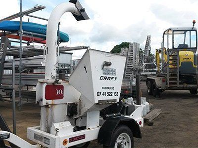 A white wood chipper on a trailer sits in an outdoor storage yard with construction equipment in the background.