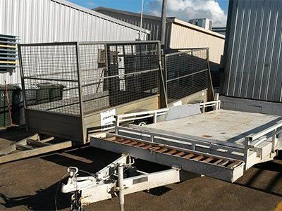 A utility trailer with a mesh caged rear section and a flatbed front, parked on asphalt near a metal building.
