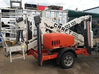 A white towable boom lift with an orange engine cover parked in an outdoor lot near several shipping containers.