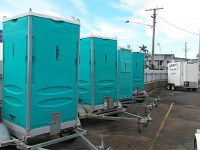 A row of teal portable toilets on trailers parked in a paved outdoor lot with a utility trailer in the background.