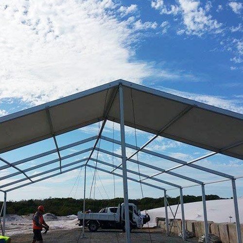 A worker stands beneath the metal frame of a large, partially assembled tent outdoors with a utility truck nearby.