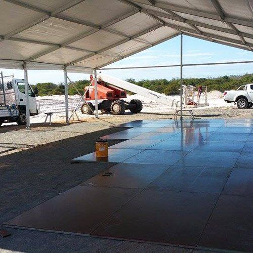 A white tent structure covers a large, dark, modular flooring system on a gravel site, with a truck and crane nearby.