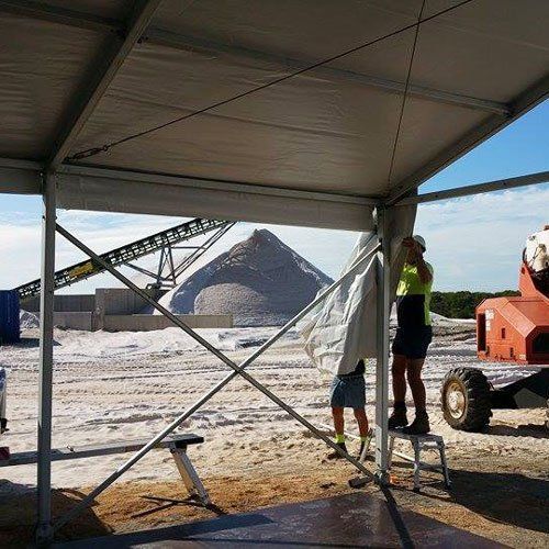 Two workers install a fabric wall under a tent frame at a large, sunlit industrial salt mining site.