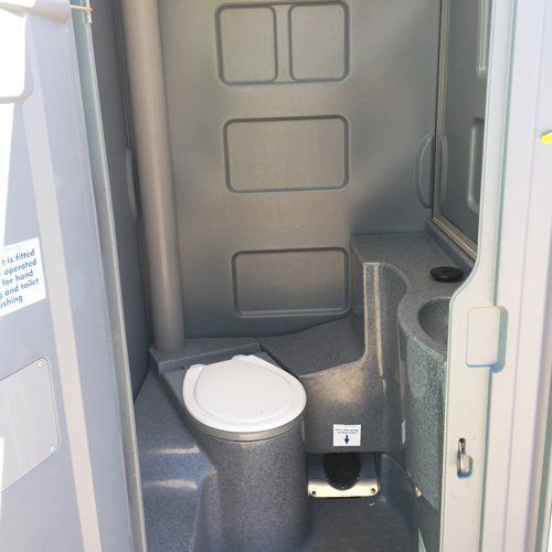 Inside a gray portable restroom with a white toilet seat, a side sink, and a foot-operated flush pedal.