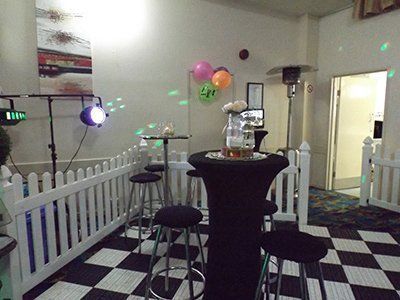 A room with a checkered floor, white picket fencing, a high table with black cover, and colorful balloons.
