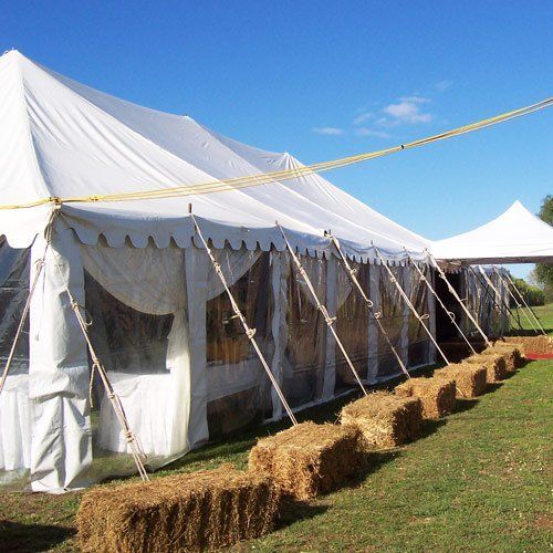A large white event tent with clear plastic sides, anchored by ropes, sitting on a grassy lawn lined with hay bales.