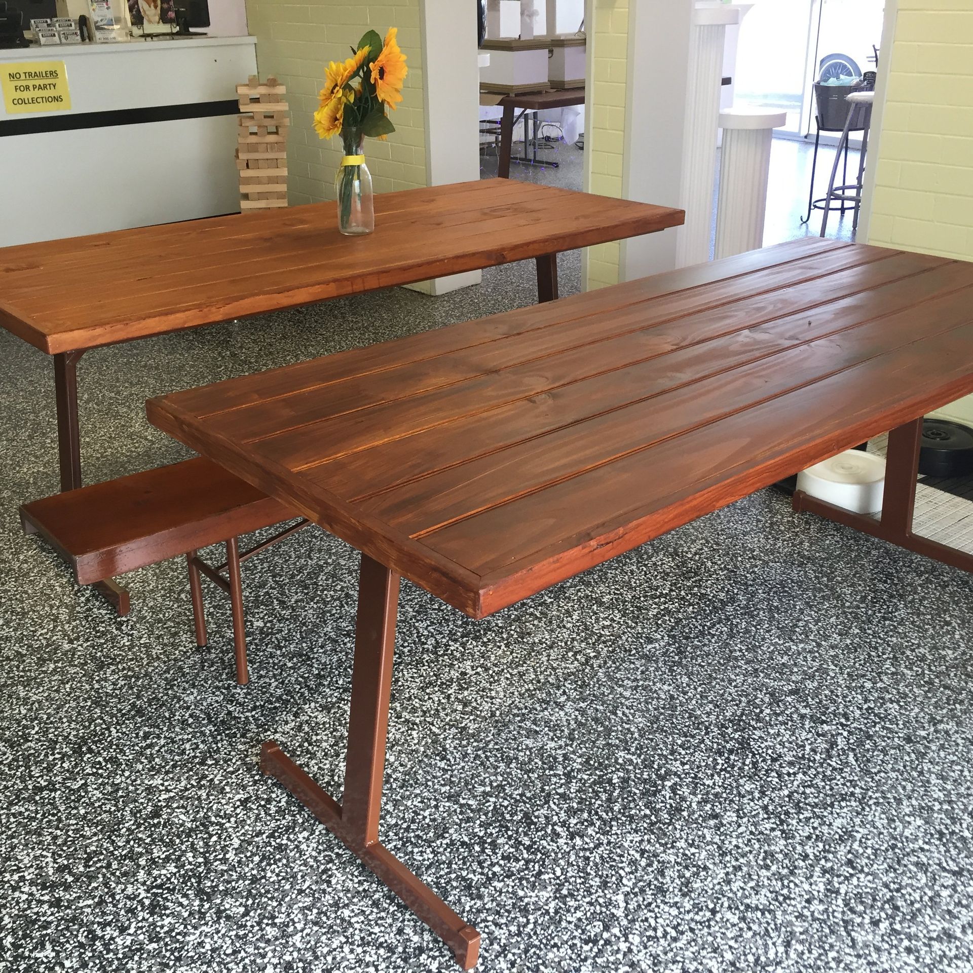 Two long, brown, wooden plank-style tables with simple metal legs stand in a room with a speckled floor.