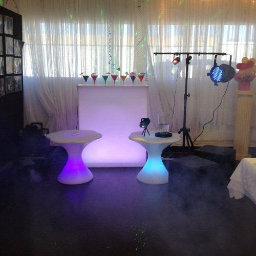 An illuminated event bar and two glowing cocktail tables in a room with white sheer curtains and stage lighting.