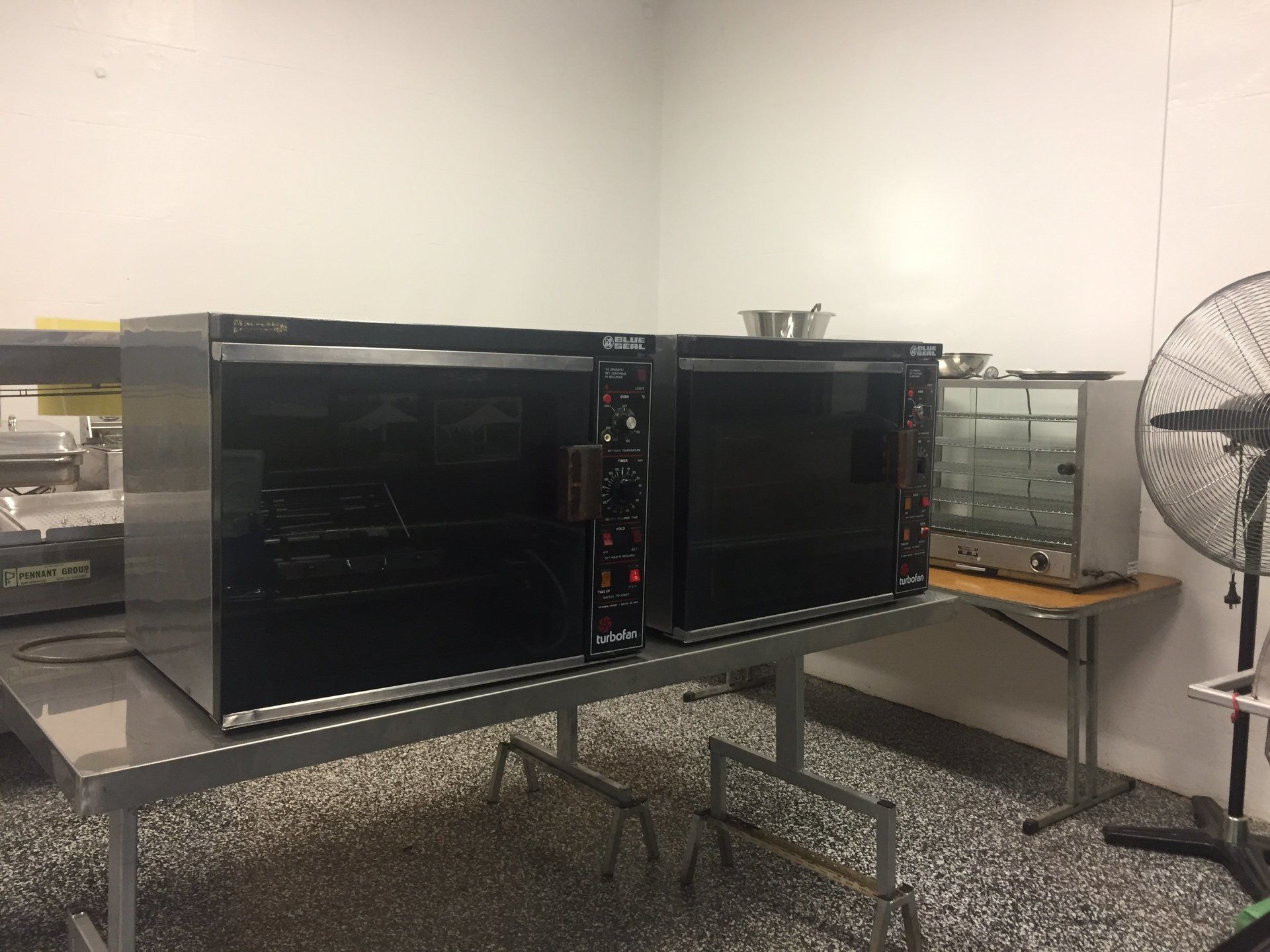 Two black commercial ovens sit side-by-side on a metal table in a kitchen, with a small warming unit and a fan nearby.