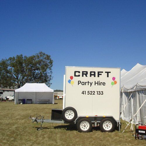 A white Party Hire trailer with balloon graphics and phone number parked on grass near white tents under a clear blue sky.