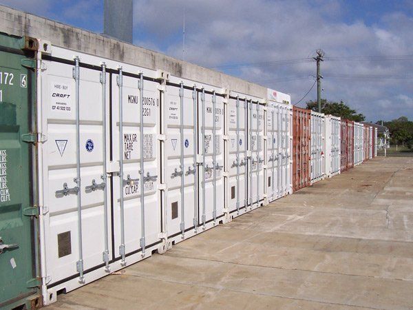 A long line of white shipping containers parked in a row on a concrete lot under a partly cloudy sky.